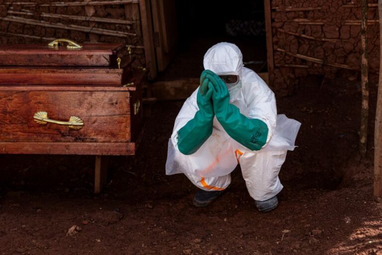 A health worker wearing protective equipment crouches beside the coffin of a suspected Ebola victim during safe burial procedures outside a family home in the community of Mongbwaluon on May 24, 2026, in Mongbwalu, Ituri Province, Democratic Republic of Congo.