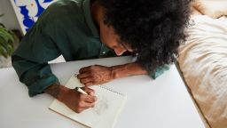 High angle of a young African American artist sitting at a table and drawing designs in a sketchbook