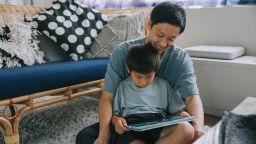 Japanese father sitting with his young son on the floor in a cozy living room, both looking at digital tablet, concept of parenting, family bonding, and digital learning. Location: indoor home setting.
