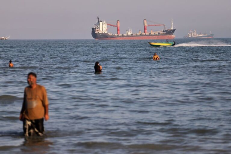 Residents swim in the waters of the Strait of Hormuz as a small motorboat passes cargo ships and other commercial vessels offshore near Bandar Abbas, Iran, Wednesday, June 17.
