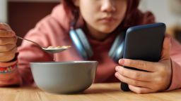 Hand of teenage girl with mobile phone having cornflakes with milk from bowl while sitting by table in front of camera and reading messages