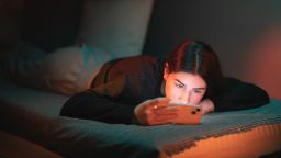 A teenage girl lies on the bed in her room lightened with orange and teal neon lights and watches a movie on her mobile phone.