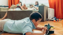 boy lying on wooden floor playing with tablet while two girls relax on sofa using smartphones with orange curtains and daylight, technology education leisure