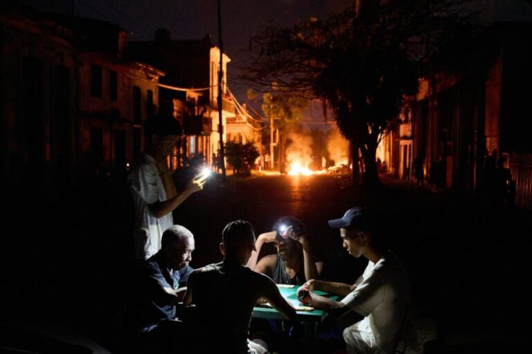 People use their phones for light while playing dominoes as a fire set by residents protesting prolonged power outages burns on a street in Havana, Cuba, on May 14.