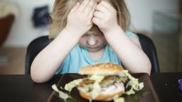 A three year old child sitting by kitchen table with a hamburger in front of her. Head resting in hands, sour face, refusing to eat her food.