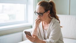 An adult woman sits on a comfortable sofa at home, holding her smartphone tightly while staring at the screen with a visibly concerned and anxious facial expression. Her furrowed brows and tense posture suggest she has just received troubling news, an upsetting message, or stressful work-related information.