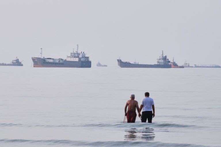 Two men wade in the waters of the Strait of Hormuz with vessels anchored in the background, off Bandar Abbas, Iran, on July 12, 2026.