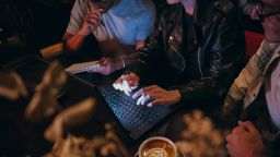 Three young adults are gathered around a table in a cozy cafe, working together on a laptop. The ambiance is warm and dimly lit, suggesting a relaxed yet focused environment as they collaborate and share ideas.