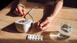 A person checking blood pressure at home using a digital monitor on a wooden table, with medication blister pack and glass of water nearby