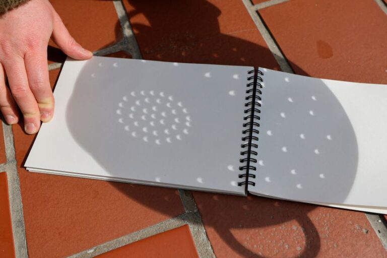 A person uses a colander and paper to see the shadow of a partial solar eclipse.