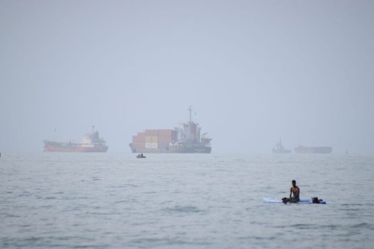 A man sits on a surfboard as a container ship and other commercial vessels appear anchored in the Strait of Hormuz off Bandar Abbas, Iran, on July 27, 2026.