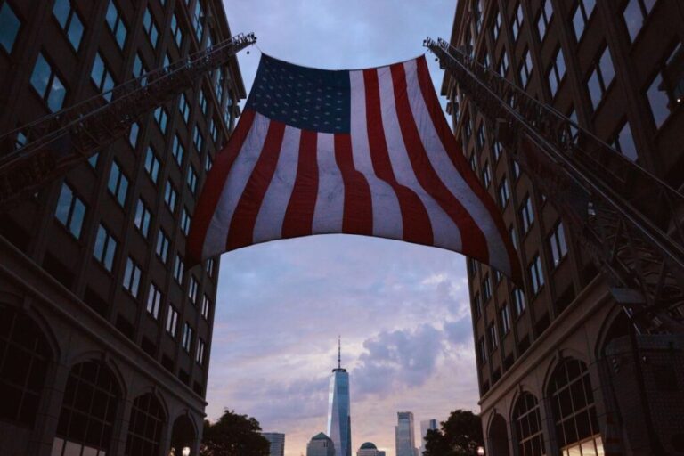 An American flag hangs between two fire trucks in New York with One World Trade Center visible in the distance.