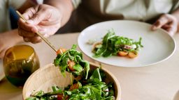 Close up on hands of Black woman putting fresh salad with green lettuce leaves on plate while having dinner at kitchen table, copy space