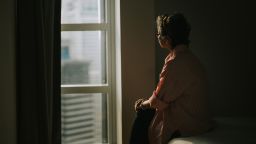 lonely asian chinese senior woman standing in the dark behind the window looking