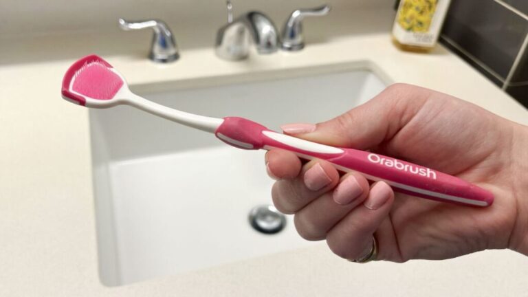 Hand holding DenTek Orabrush Tongue Cleaner in front of bathroom sink.